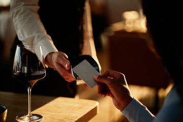 Restaurant worker holding payment terminal while client handing credit card for payment in restaurant setting, wine glass visible on table