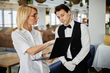 Caucasian middle aged woman showing laptop screen to Caucasian young adult man in waiter uniform...