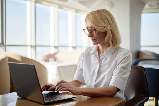 Caucasian middle aged woman working on laptop in restaurant setting, sitting at table and typing on keyboard, focused on screen, wearing glasses, natural light coming through windows