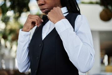 Black young adult woman adjusting bow tie while preparing for restaurant shift, wearing formal...