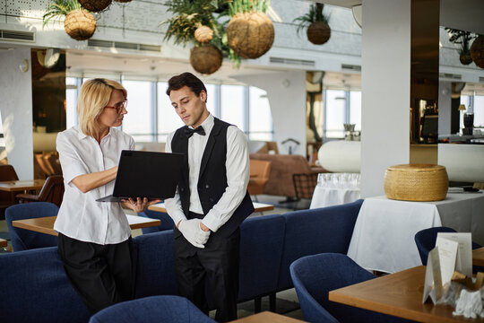 Caucasian middle aged woman showing information on laptop to Caucasian young adult man in waiter uniform standing together in modern restaurant during work discussion