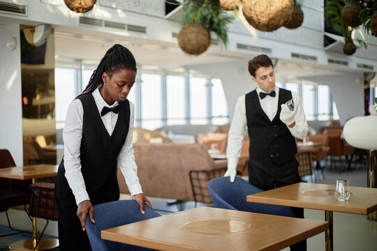 Black young adult woman arranging chair while Caucasian young adult man standing nearby in modern restaurant interior, both wearing formal uniforms and focused on tasks