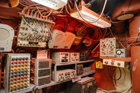 An array of analog controls, wires, and switches fills a submarine communication room. A warning sign, colored buttons, and monitoring devices are mounted across red metal walls.
