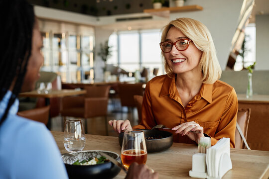 Caucasian middle aged woman smiling and talking with Black woman while sitting at restaurant table, both women eating and drinking, engaging in friendly conversation during meal
