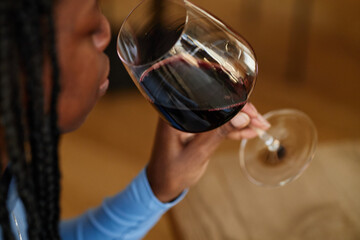 Black woman holding wine glass near lips sitting at restaurant table drinking red wine closeup showing partial face and hand