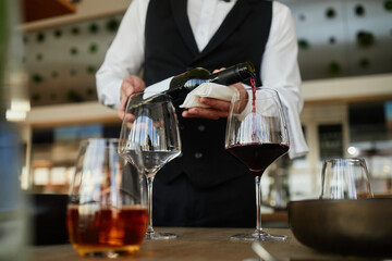 Unrecognizable waiter pouring red wine into glass, wearing formal vest, standing behind restaurant counter, hands visible holding bottle and napkin, focus on wine service