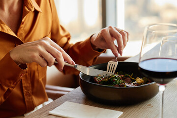 Woman eating meal with fork and knife at restaurant table, hands visible holding utensils, glass of red wine on table, natural daylight coming through window