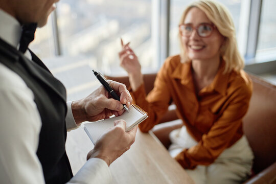 Caucasian young adult woman smiling and gesturing while sitting at restaurant table, male waiter taking order and writing in notepad, both interacting during service - Powered by Adobe