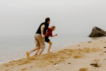 Young caucasian couple running and laughing on sandy beach by the ocean