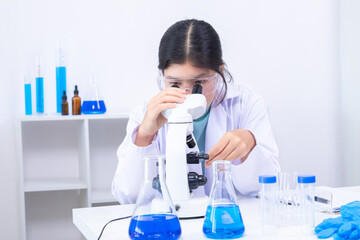 A young Asian female scientist in lab coat observes samples through a microscope in a clean science...