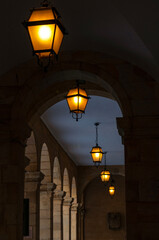 Arcades, skyline with European architecture and lit street lamps. A night walk under the arches in a deserted city, dark alley lit by street lamps. Symbolic, mystery, crime, thriller, spy story