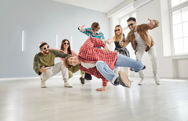 Happy young man performing impressive breakdance move in front of cheering friends in modern studio. Joyful group of hip hop dancers having fun and showing support during energetic dance session.