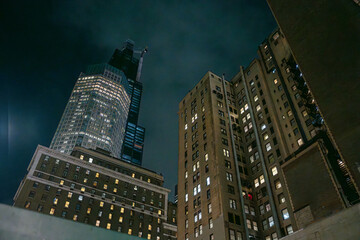 Tall buildings in New York City rise into a dark sky, glowing with window lights. A modern skyscraper under construction stands beside older brick architecture, creating a dramatic night contrast.
