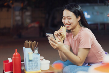 Young woman enjoying street food while using smartphone in Hanoi, Vietnam at night