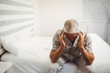 Senior man sitting on bed edge wearing plaid pajamas pressing temples in room with sheer curtains