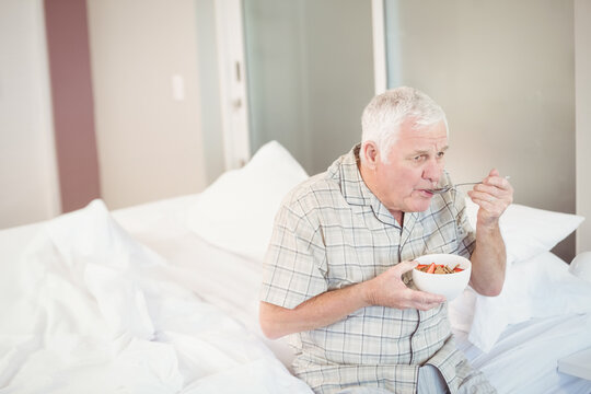 Senior man sitting on bed scooping cereal with metal spoon from white bowl beside open door