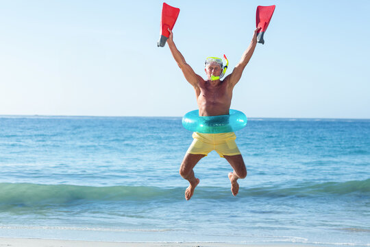 Senior man jumping midair on sandy shore, wearing snorkel mask with green ring and red fins