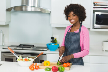Knife is slicing green bell pepper on wooden cutting board in bright kitchen, with salad bowl