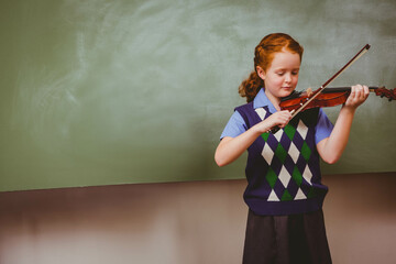 Schoolgirl playing violin with bow before green chalkboard in classroom wearing uniform, copy space