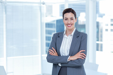 Woman in grey blazer striped blouse standing with arms crossed at office windows city view
