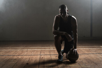 Male athlete kneeling on lined court holding basketball under lighting and smoke drifting by beam