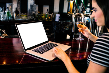 Woman holding glass of white wine and typing on open laptop at polished wood bar counter