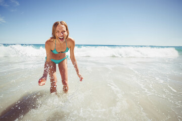Woman bending and laughing while splashing shallow ocean waves at sandy beach under clear sky