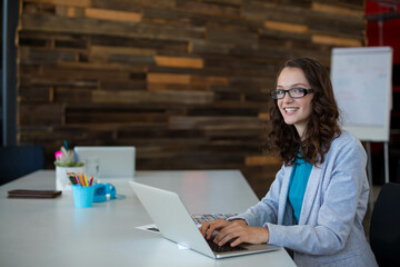 Woman in teal blouse, glasses typing on laptop at coworking table with notebook, copy space