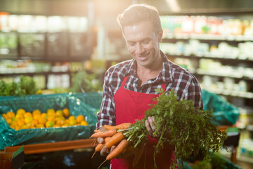 Man in produce section wearing red apron holding carrots and inspecting wooden bin with oranges