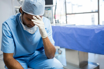 Asian male surgeon sitting on stool wearing scrubs, mask, gloves in operating room, copy space