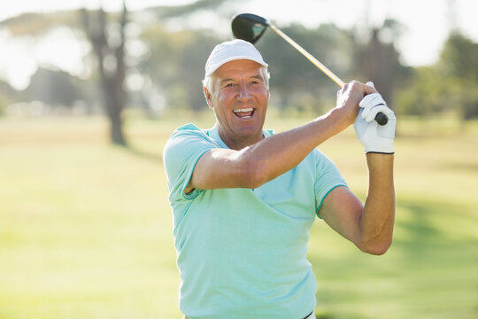 Senior man swinging driver and smiling finishing golf swing on fairway in polo shirt and glove