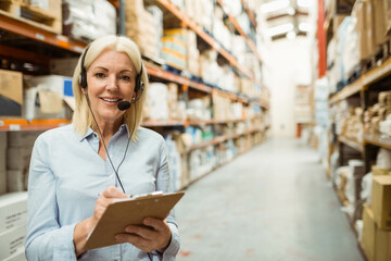 Woman checking inventory on clipboard while wearing headset among metal racks in warehouse aisle