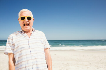 Senior man standing on sandy shore at beach wearing striped shirt and yellow sunglasses, copy space