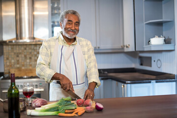 Senior African American man chopping vegetables on wooden cutting board in kitchen with wine bottle