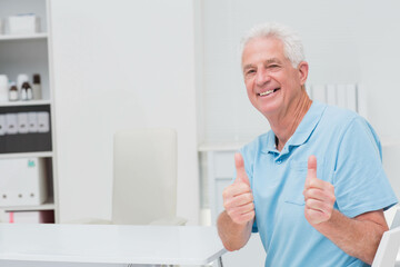 Senior man smiling, giving two thumbs up in clinic with white desk, medical bottles, copy space