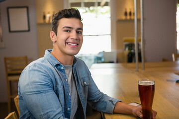 Man sitting at pub counter wearing chambray shirt sipping beer from pint glass with smartphone