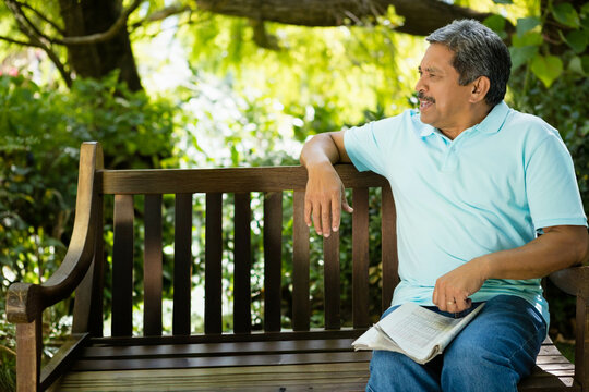Senior man sitting on wooden bench in shaded park holding folded newspaper, copy space - Powered by Adobe