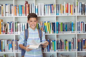 School-age boy standing in school library wearing backpack reading open book before white bookshelf