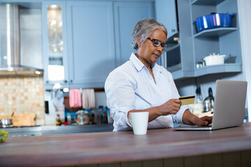 Senior African American woman checking credit card at kitchen counter using laptop with white mug