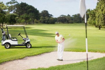 Senior man taking bunker shot hitting golf ball toward flagstick in sand trap, copy space