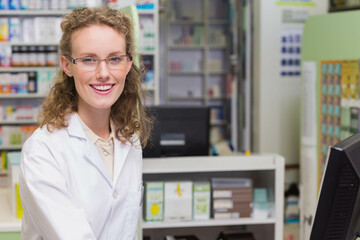 Smiling female pharmacist in lab coat standing behind pharmacy counter using computer and register
