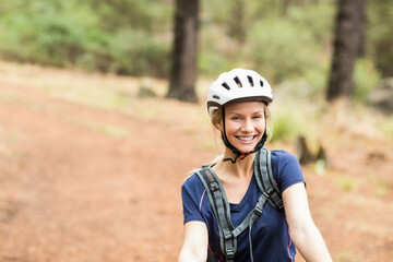 Woman riding bicycle wearing white helmet and gray hydration pack on red dirt forest trail