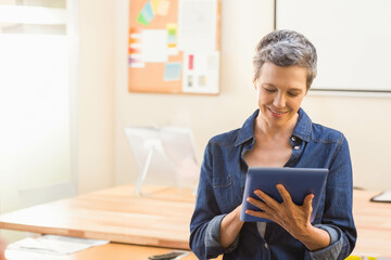 Mature woman holding tablet and smiling by wooden table with monitor, boards at office, copy space