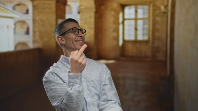 Caucasian man outdoors at an old university smiling mischievously while making an inappropriate hand gesture emphasizing unconventional expression under sunny conditions.