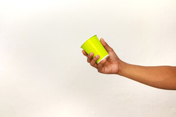 A brown-skinned hand holds a vibrant green disposable cup against a white background, providing a...