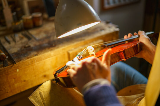 Violinmaker meticulously polishing wooden violin body, working under bright lamp surrounded by crafting tools in traditional workshop setting
