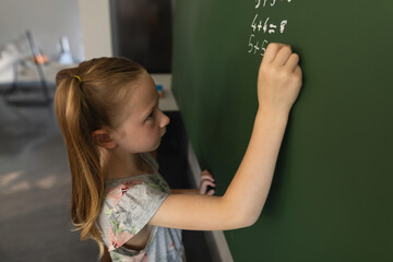 Student writing math equations with white chalk on green chalkboard, desks and chairs in background