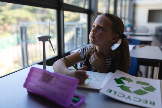 Girl is sketching with green pencil by window gazing outside by wind turbine model, recycling sheet