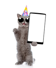 Laughing cat with open mouth wearing sunglasses and party cap standing on hind legs and showing big smartphone with blank screen. Empty space for text. isolated on white background