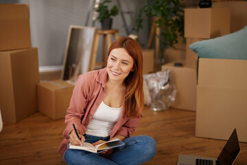 In a new home, a woman with red hair sits on the floor among cardboard boxes. She writes in her...